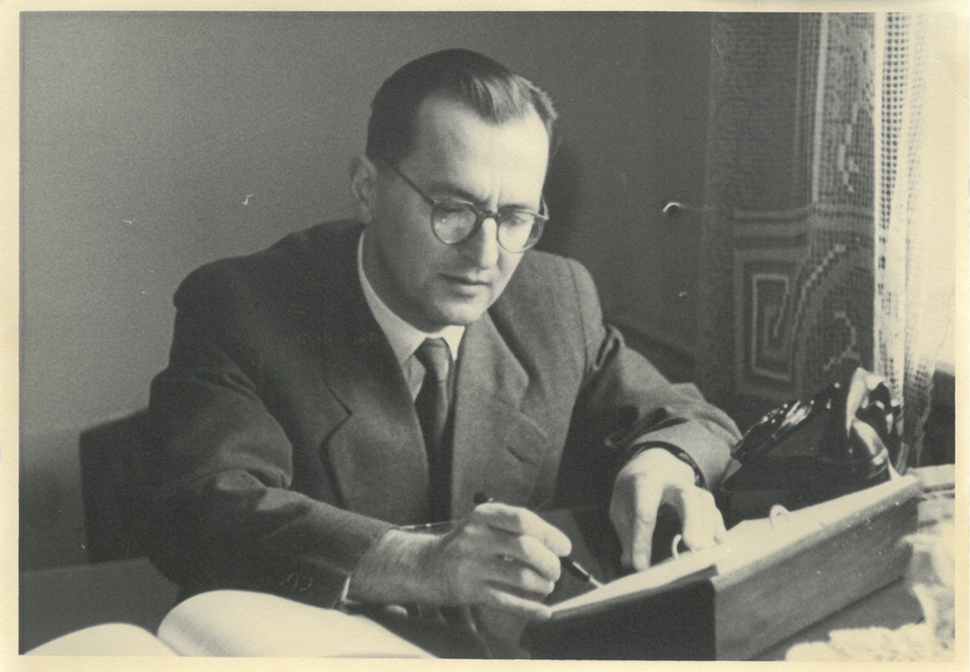 Czarno-białe zdjęcie. Mężczyzna siedzący przed biurkiem nad dokumentami.